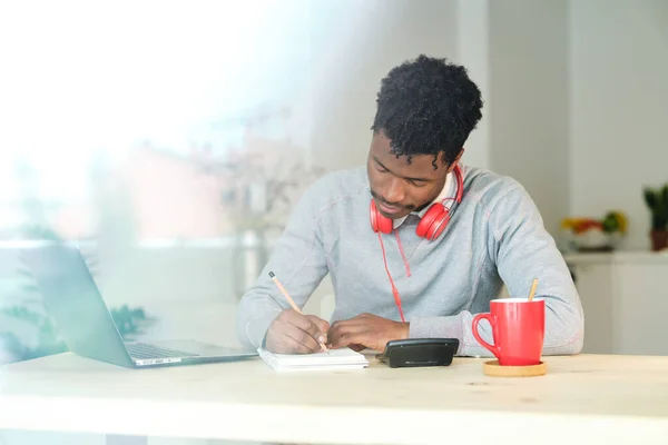 Black man writing with pencil in notepad while doing homework at desk with calculator and cup of hot drink at home