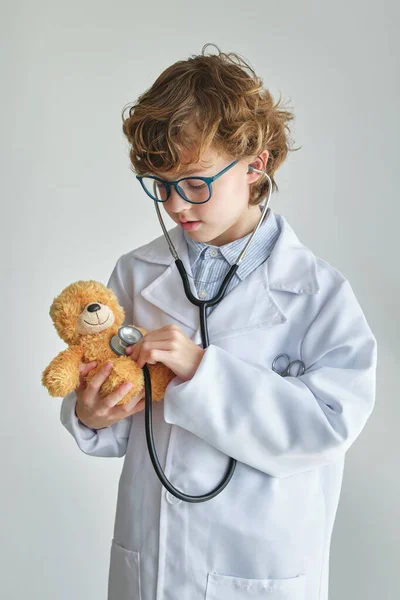 Child in eyeglasses and medical robe listening to heart rate of toy bear with stethoscope on white background