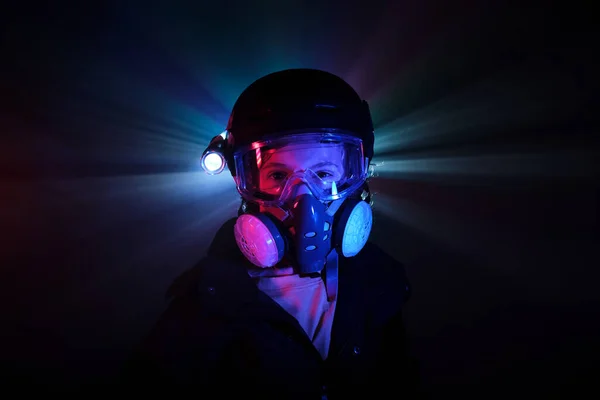 Boy in protective respirator with shining flashlight looking at camera while standing in dark mint with neon lights during illegal heist