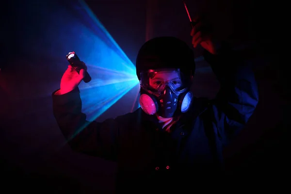 Child in gas mask with filters shining with flashlight while standing in darkness during robbery in mint against blue neon light