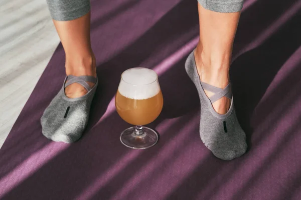 From above of crop unrecognizable woman in gymnastic slippers standing on yoga mat near brandy snifter with ale in sunny studio