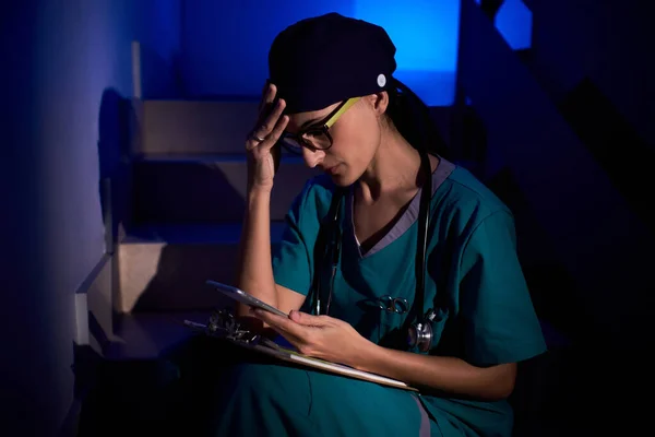 Confused female doctor in medical uniform with stethoscope sitting on staircase with clipboard and browsing mobile phone in darkness under blue illumination