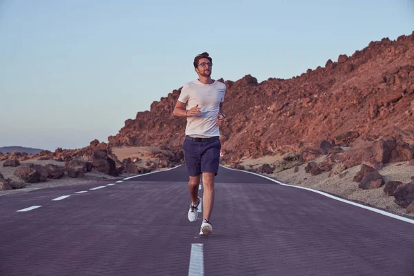 Full body of active male runner in activewear running on asphalt highway near rocky mountain range in nature on summer day