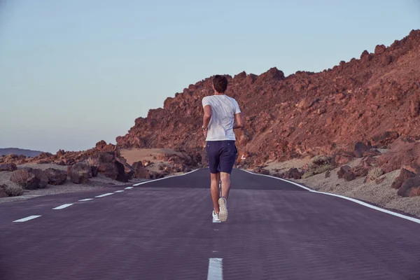 Full body back view of anonymous active male running on empty asphalt highway near mountain ridge in nature on summer day