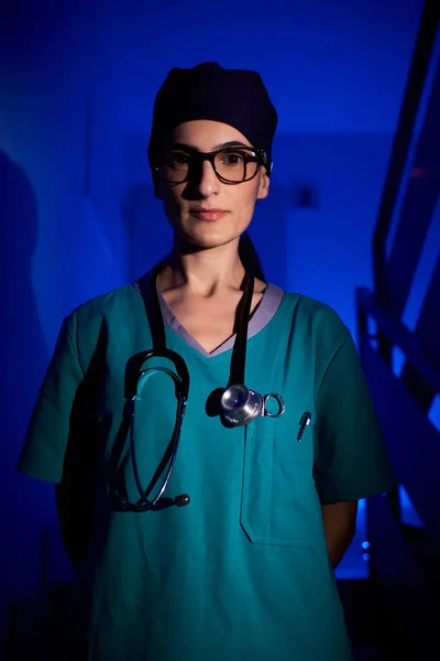 Confident adult woman in medical uniform with stethoscope standing on stairs with hands behind back and looking at camera in dark room under blue illumination