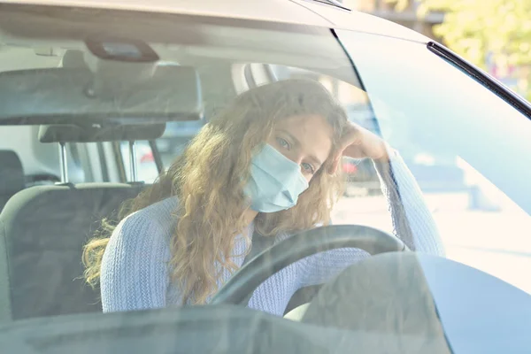 Thoughtful young female driver in protective face mask sitting in car drivers seat and leaning on hand while looking at camera on sunny summer weather