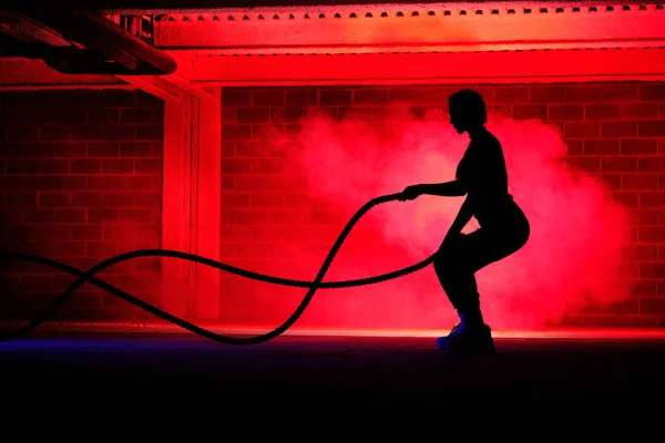 Side view of unrecognizable sporty lady standing near brick wall and performing exercise with heavy ropes in red neon illumination