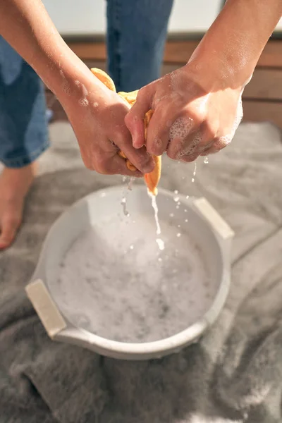 From above of faceless person in jeans standing on towel and wringing out rag over white basin with foamy water while doing household routine at home