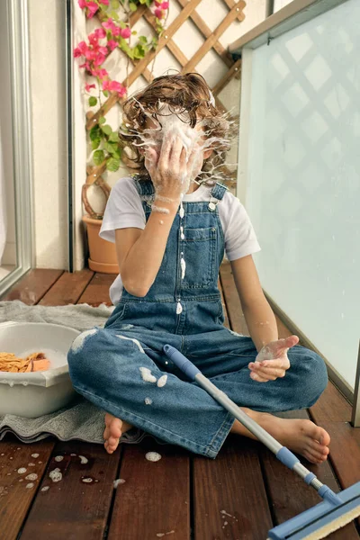 Barefoot anonymous preteen boy sitting on wooden balcony floor and hitting face with foam splashes while washing glass in sunlight