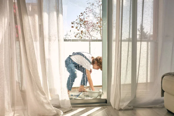 Side view of boy with curly hair standing on towel and rinsing rag in basin while doing household chores and washing windows on balcony at home