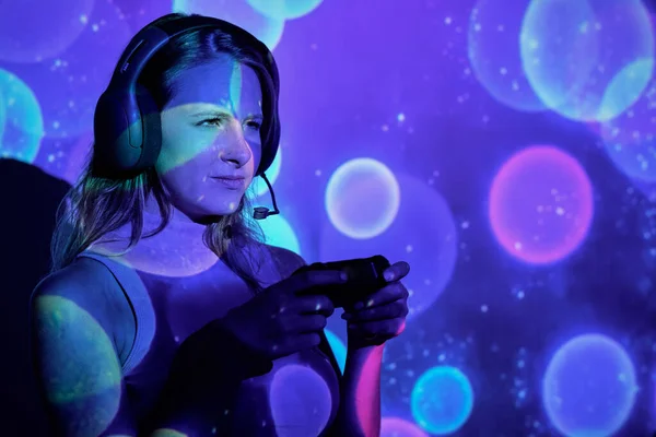 Dissatisfied young woman frowning and looking away while using gamepad to play challenging videogame under colorful sparkles projection