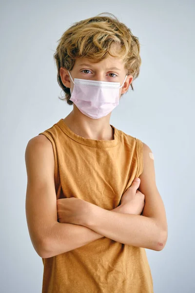 Self confident child in undershirt and disposable mask looking at camera with folded arms on light background
