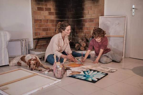 Full body of young female artist in shirt having fun with curly haired kid and painting abstract picture together on paper on floor in country house