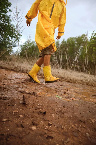 Unrecognizable child in yellow raincoat and gumboots playing in puddle while walking in nature on rainy day