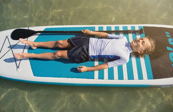 From above full body of happy boy in activewear lying on paddleboard and relaxing in middle of sea during summer vacation