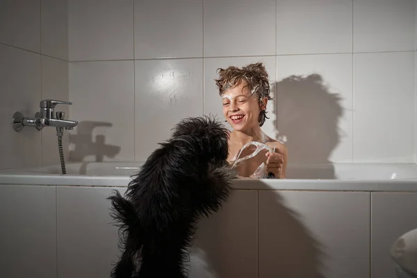 Delighted boy covered with foam sitting in bathtub near cute black dog during daily routine in light bathroom at home