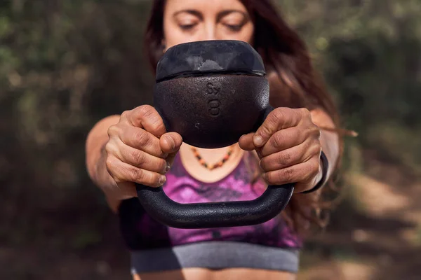Crop female in sports top lifting heavy kettlebell during training with weight for strengthening muscles at sunlight in woods