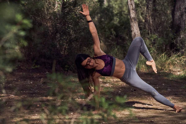 Full body of barefoot Hispanic female meditating with closed eyes while doing Side Plank Star pose for improving sense of balance and focus during yoga practice in woods