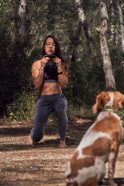 Full body of Hispanic female athlete in sport clothes exercising with kettlebell near dog on path against trees in daylight