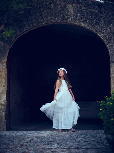 Full body side view of young bride in white dress and floral wreath looking away while standing on old stone wall entrance