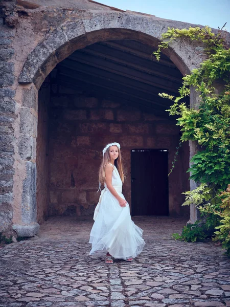 Full body of charming adolescent with long hair wearing lace bridal gown standing near arched entrance to ancient stone house