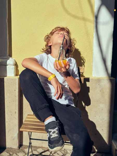 Cute boy in casual wear drinking fresh juice with head thrown back while sitting on sunny street near building in city