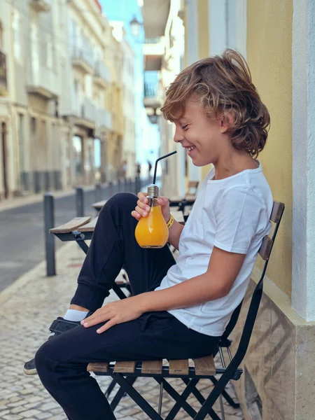 Side view of positive boy drinking refreshing juice from bulb shaped bottle while sitting on street near building in city