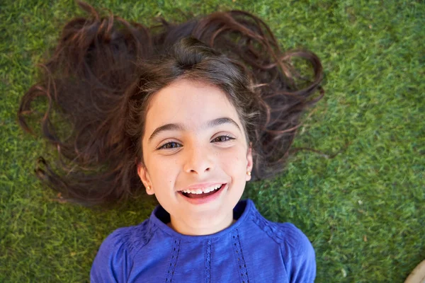 Top view of happy child with brown eyes resting on green lawn and looking at camera on weekend summer day