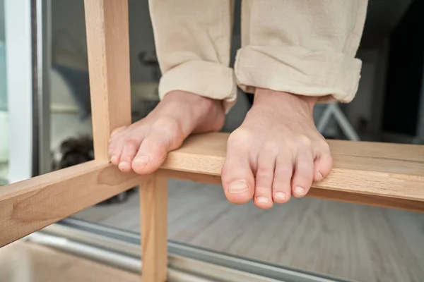 Bare feet of unrecognizable person in light pants standing on wooden table at daytime at home