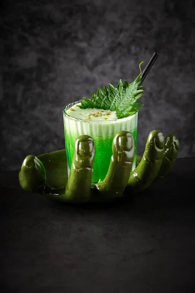Glass of refreshing green cocktail with whipped foam decorated with shiso leaf and grated camomile flower on serving plate in form of green hand in restaurant