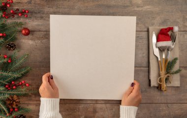 Bir kadın Noel yemeği masasında otururken menüyü elinde tutuyor. Noel ağacı süslemeleri, eski ahşap masa üzerinde çatal bıçak takımı ve yılbaşı menüsü ile sınırdır..