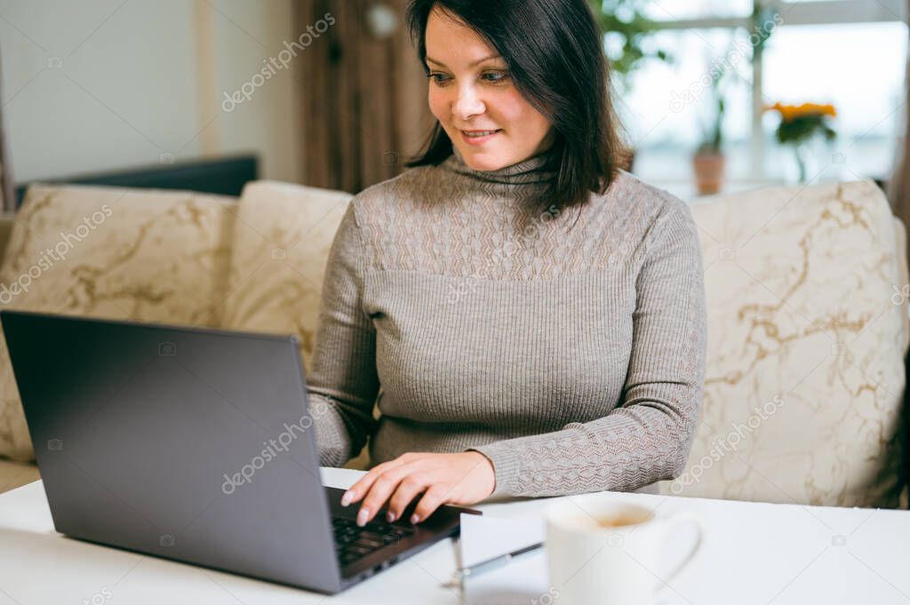 woman works on a laptop remotely on the background of the interior of the living room. Businesswoman 30-40 years old sits on couch at home and works on laptop. Female freelancer with hands on keyboard