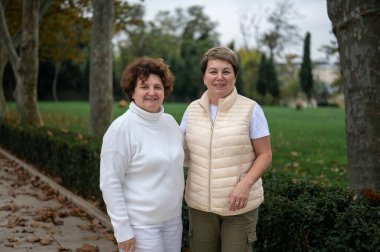 Dışarıda durup kameraya bakan iki yaşlı kadın. İki orta yaşlı kadın parkta yürüyor sonbahar serin havası