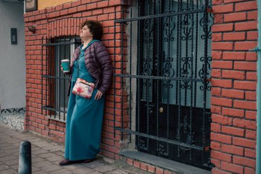 Elinde kahve, tuğla duvara yaslanmış yaşlı bir kadın, hayatı gözleri kapalı düşünüyor.