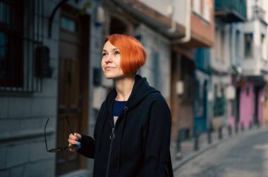 Genç lezbiyen kadın güvenle caddede yürüyor ve önüne bakıyor. Dar yolda parlak kızıl saçlı neşeli genç bir kadın yürüyor. Çekici kadın dar şehir caddesinde yürüyor