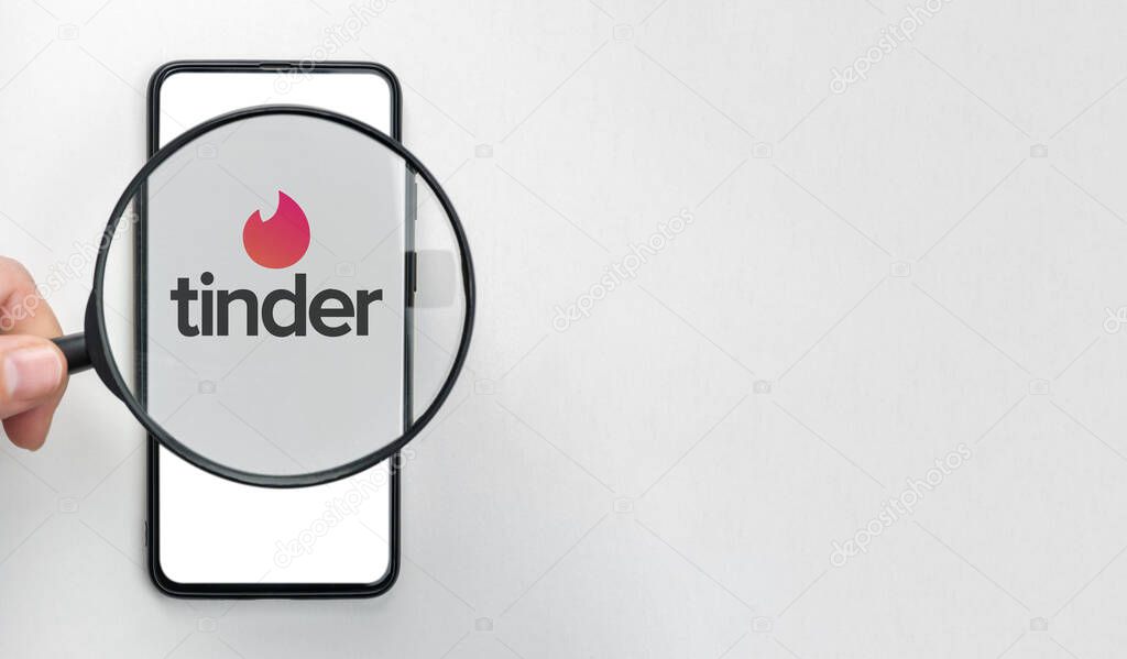 New York, USA - January 11 2022: Tinder dating app logo under a magnifying glass. Top view on white table. Tinder is a popular designed for romantic Dating in accordance with parameters geolocation