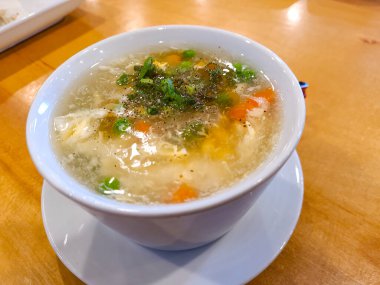 Tahta bir masada servis edilen beyaz bir kasede soğanlı, bezelyeli ve havuçlu yumurta çorbası.