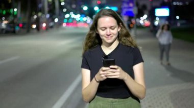 Geceleri sokakta yürürken telefonuyla mesajlaşan genç bir kadın.