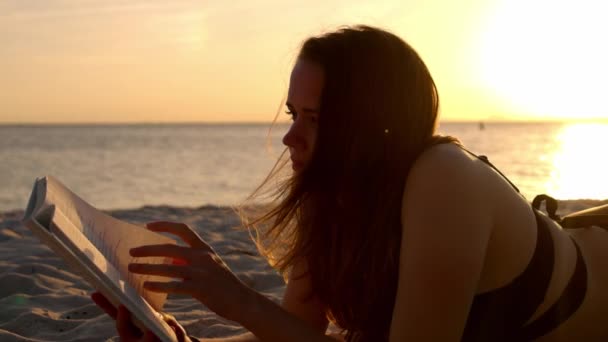 Jovem lê um livro enquanto está deitado na praia ao pôr do sol — Vídeo de Stock