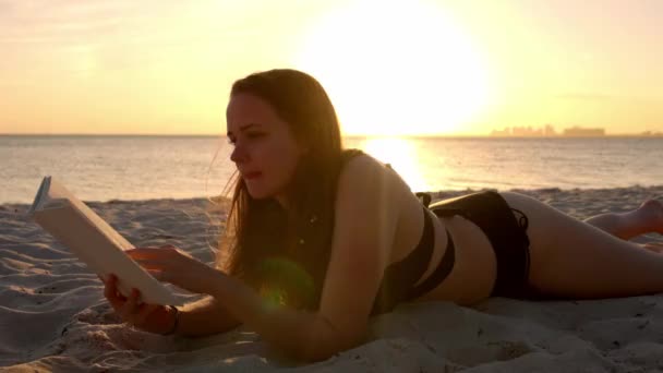 Mladá žena čte knihu, zatímco leží na pláži při západu slunce — Stock video