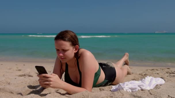 Ładna kobieta relaksuje się na plaży SMS-ując komórką — Wideo stockowe