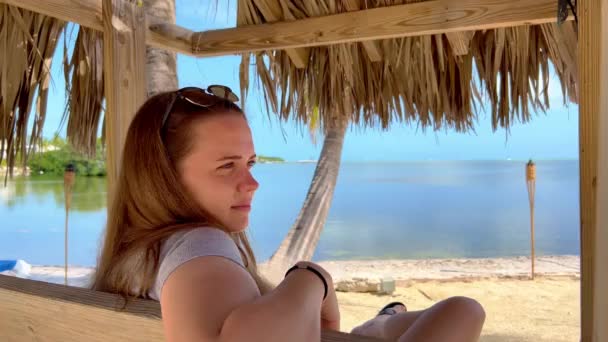 Райський пляж - молода жінка, яка сидить на лаві, насолоджуючись атмосферою карібів. — стокове відео