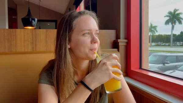Mulher jovem senta-se em um restaurante e bebe suco de laranja — Fotografia de Stock