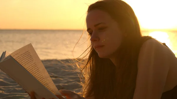 Jovem lê um livro enquanto está deitado na praia ao pôr do sol — Fotografia de Stock