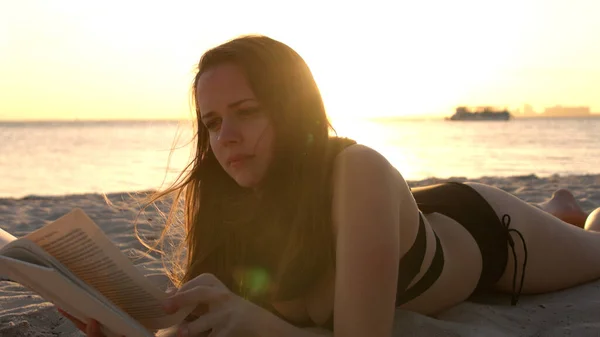 Mujer joven lee un libro mientras está acostada en la playa al atardecer — Foto de Stock