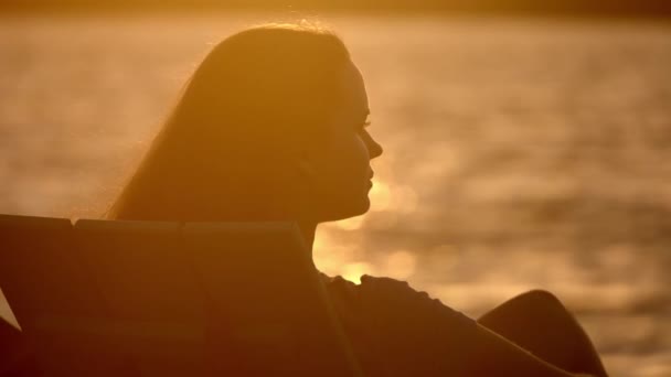 Νεαρή γυναίκα κάθεται στην παραλία και απολαμβάνει το ηλιοβασίλεμα — Αρχείο Βίντεο