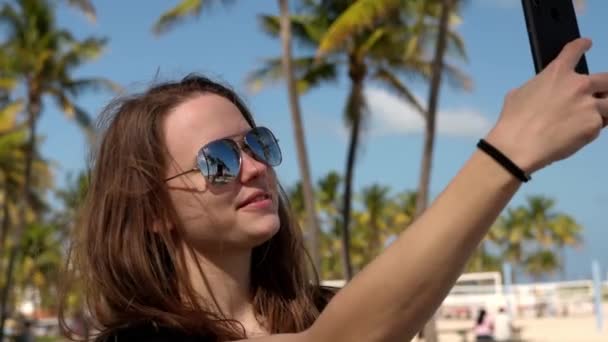 Young woman taking a selfie under palm trees at the beach — Stock Video