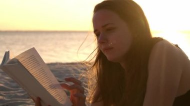 Genç kadın gün batımında sahilde yatarken bir kitap okuyor.