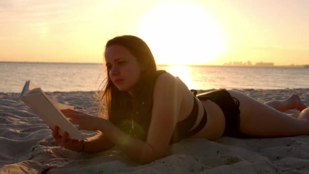 Young woman reads a book while lying on the beach at sunset — Stock Video
