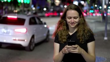 Geceleri cep telefonuyla mesajlaşırken sokakta yürüyen mutlu genç bir kadın.